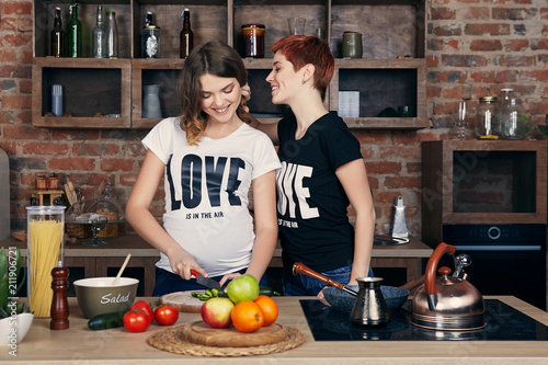Lesbian pregnancy. Two young healthy women, one of them pregnant, enjoying togetherness in the kitchen. The beautiful expectant mom cutting vegetables while her girlfriend touching her hair tenderly.