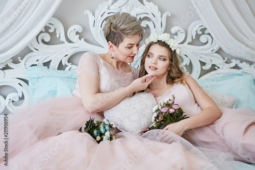 Lesbian honeymoon. Newlyweds in light pink bridal dresses lying on a luxury bed and enjoying togetherness, with bouquets in their hands. The short-haired bride touching tenderly her spouse's chin. photo