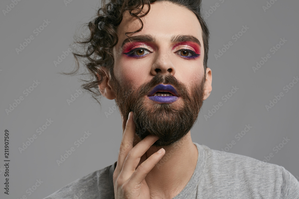 Male makeup look. Closeup portrait of a young bearded man, wearing purple  lipstick, winged eyeliner, pink