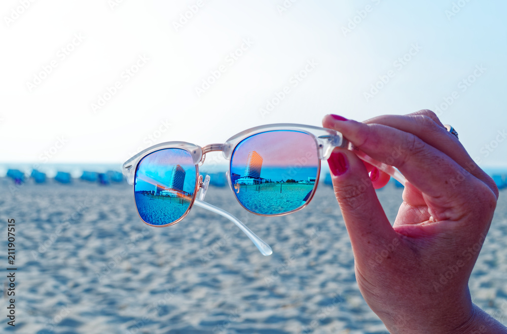 Sonnenbrille mit dem Hotel Neptun in Warnemünde im Hintergrund am Strand im Sonnenschein von einer Hand gehalten - obrazy, fototapety, plakaty 