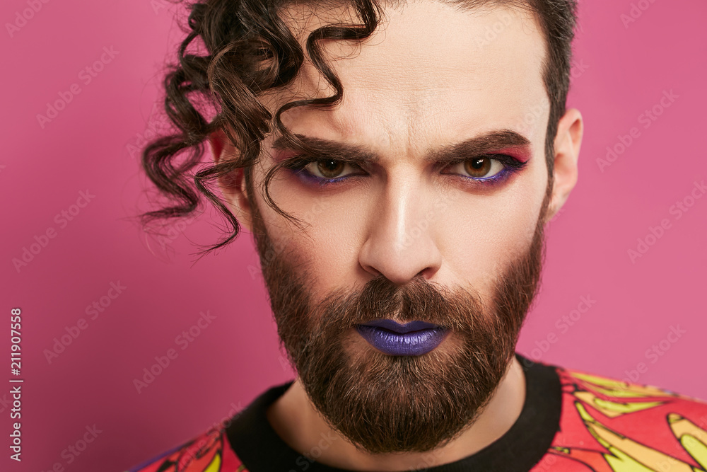 Male look. Closeup portrait of a young man, rocking hipster beard, curly hairstyle, purple lipstick, winged eyeliner, pink eyeshadow. The guy looking at the camera over pink background. Stock