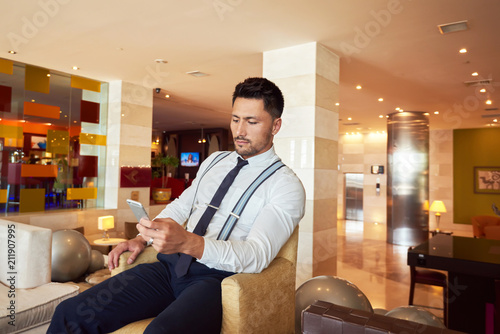 Worried businessman is reading e-mail on mobile phone from investor about suppression of the financing of his project, while is sitting in the lobby of an expensive hotel photo