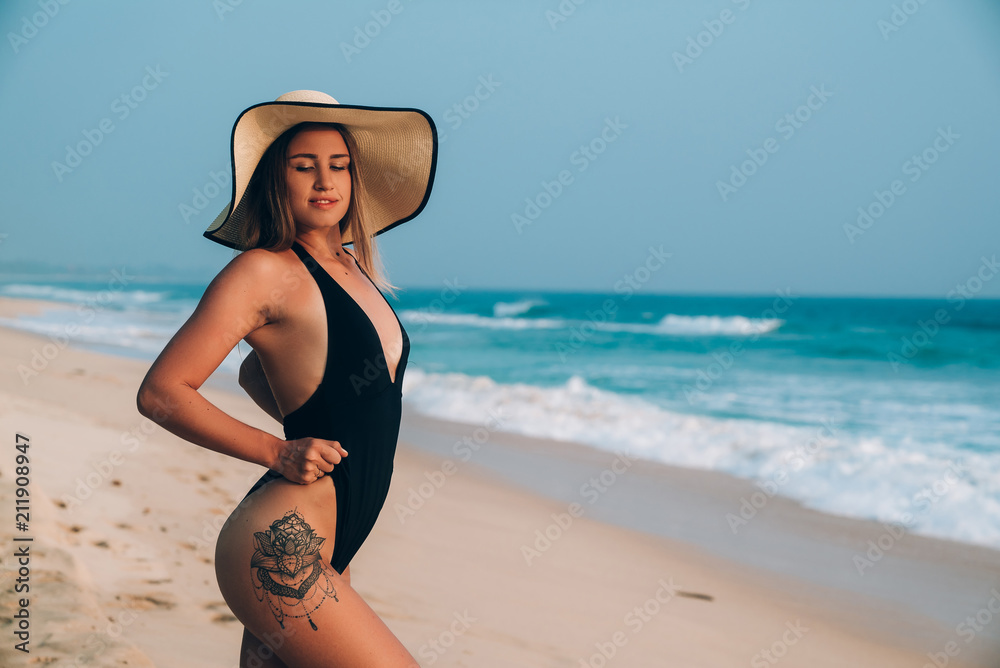 A slender European woman poses in a fashionable light hat and a black swimsuit, shows her hips with a tattoo, has a round ass