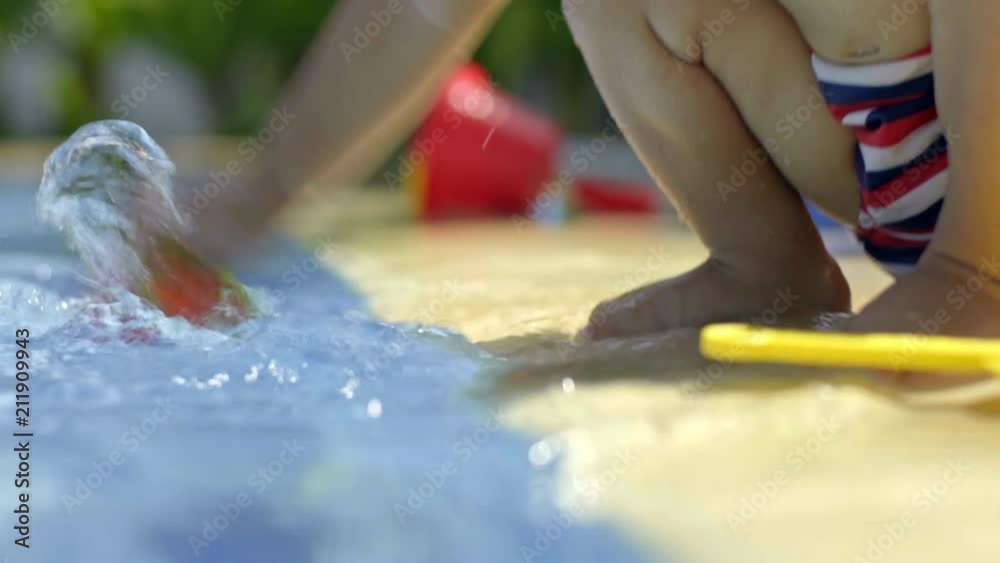 Adorable baby boy playing with toy plastic cups and splashing water in swimming pool at sunny day outdoors