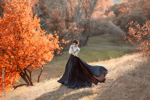 The lady in a vintage dress strolls along the picturesque, golden, autumn hills at sunset. A long train of black skirt fluttering in the wind. The girl is hugging herself from the cold. Art Photo