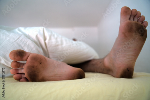 Dirty bare feet of a sleeping person showing out of the blanket on a bed