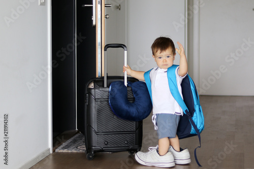 Foto Cute little baby boy in fathers sneakers and with big backpack holding suitcase and waving goodbye staying at open door ready to travel