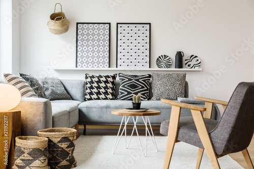 Real photo of a small table standing between a sofa with pillows and an armchair on a white rug in boho living room interior with posters on a shelf