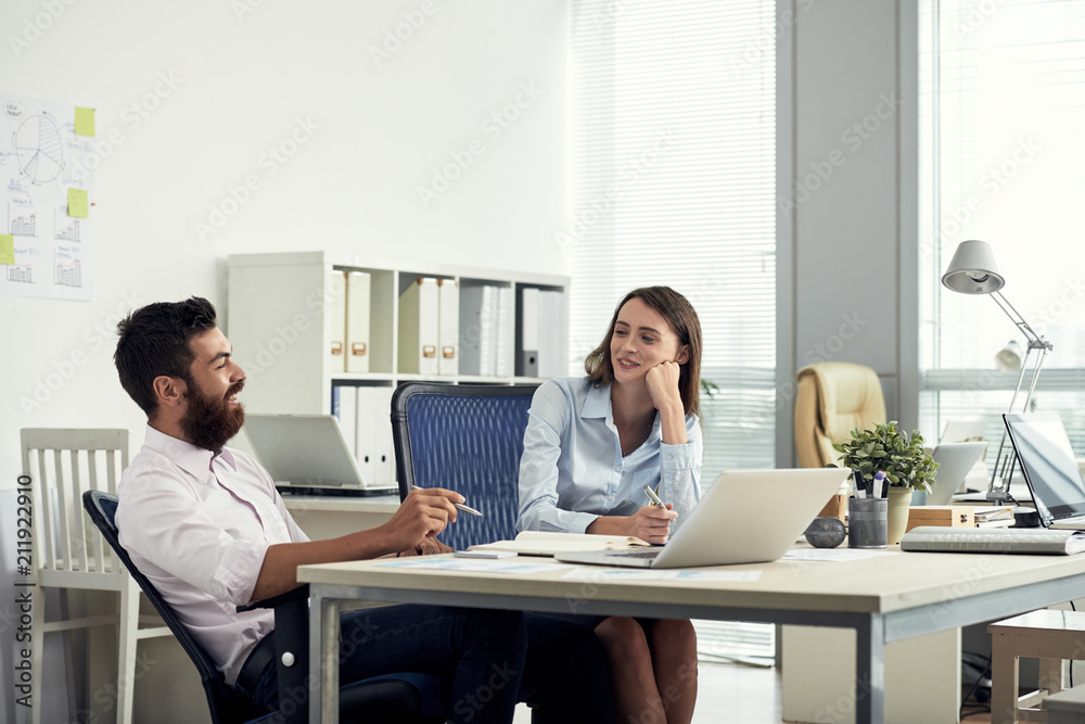 Young bearded man and elegant woman sitting at table in office and having rest while chatting cheerfully