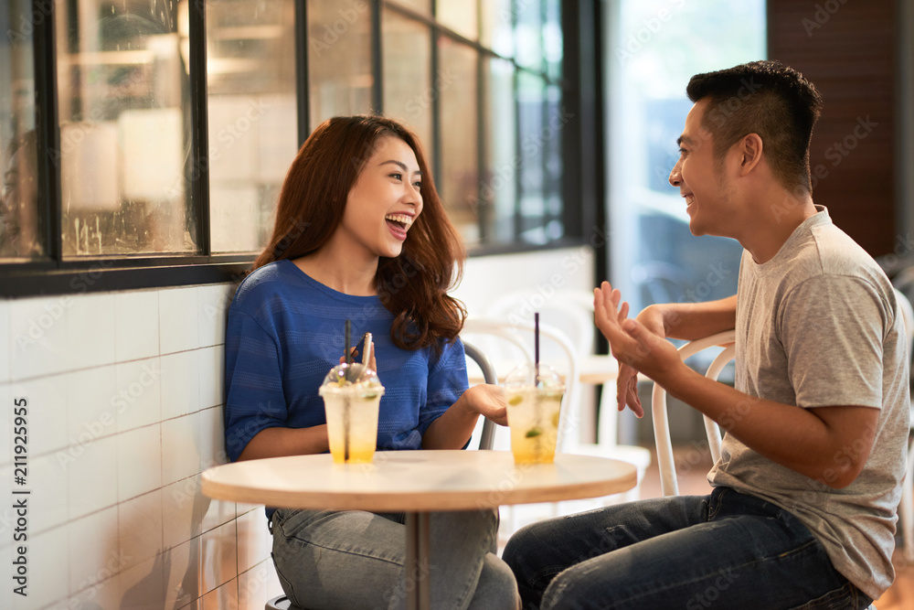 © DragonImages - Asian young couple chilling in cafe having drinks in cups and laughing happily while chatting