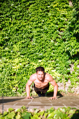 Determined shirtless young man doing push-ups. Front view, lush green foliage in background