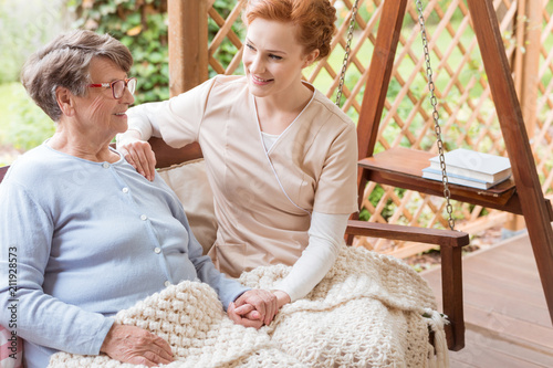 Obraz na plátně Young volunteer and a senior woman sitting together on a garden swing, holding h