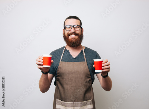 Happy bearded barista wearing apron and glasses on white background, holding two red paper cups. Hipster man smiling at the camera.