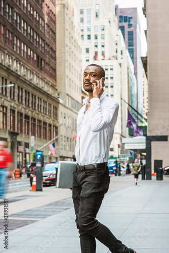 Young African American businessman with beard talking on cell phone, traveling in New York, wearing white shirt, carrying laptop computer, walking on street in Manhattan. Cars, buildings on background