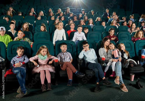 Front view of many children and teenagers sitting in movie theatre and enjoying cartoon. Cute little girl sitting on her mother laps. Spectators expressing different emotions. Project light source.