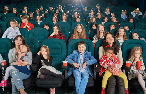 Young spectators sitting in modern movie theatre and watching cartoon. Teenagers and children sipping fizzy drinks and eating popcorn. Cute girls sitting on their moms laps. Project light source.