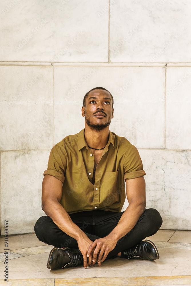 Young African American Man with beard, wearing green short sleeve shirt, black pants, leather shoes. legs crossed, sitting on marble ground by wall on campus in New York, raising head, looking up..