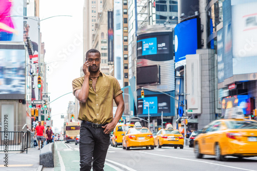 Young African American Man traveling in New York, wearing green short sleeve shirt, walking on busy street in Times Square of Manhattan, talking on cell phone. High buildings, cars on background..