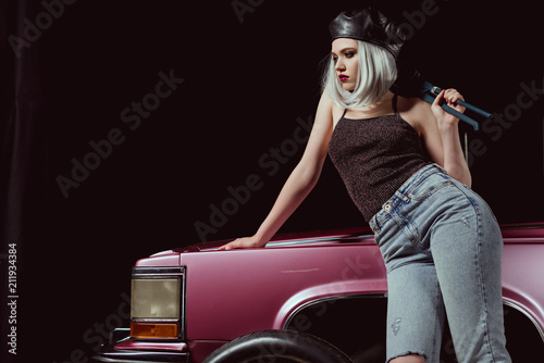 stylish blonde woman in beret leaning at retro car and looking away on black
