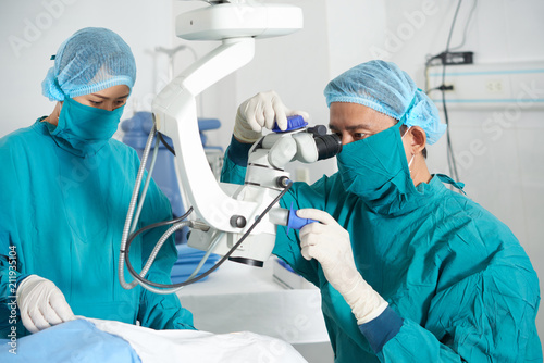 Focused Asian man looking at operating microscope while female colleague preparing patient for surgery