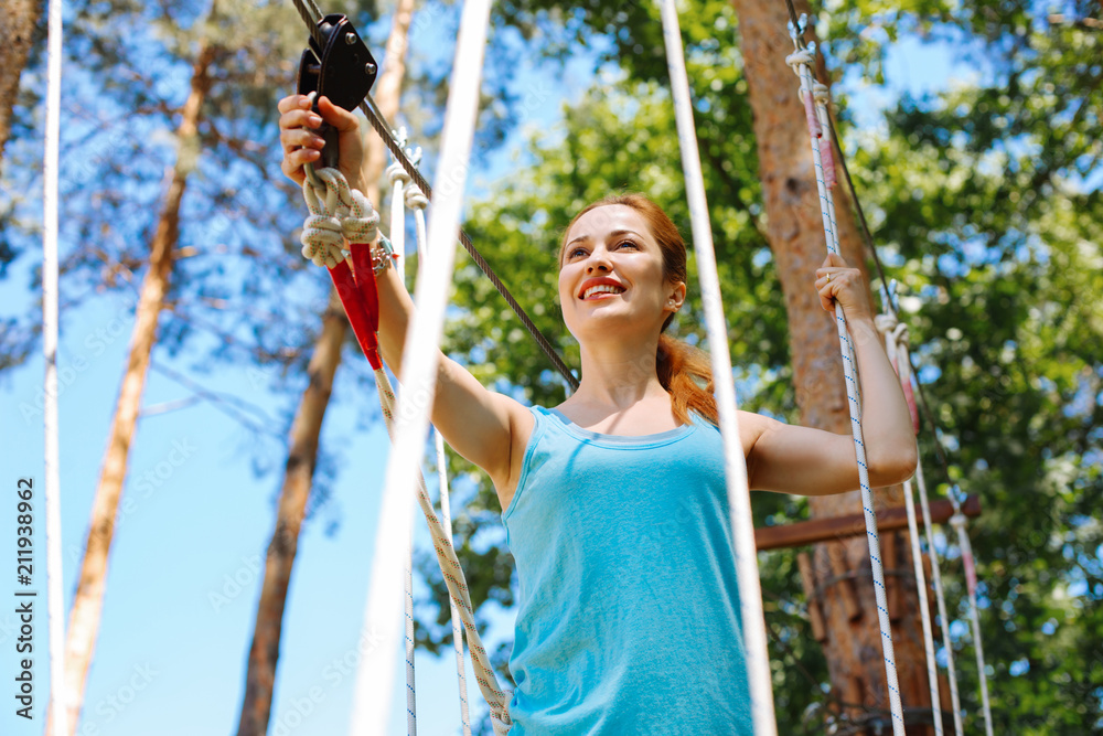 Enjoying extreme experience. Cheerful young woman climbing at a rope park and smiling happily while having a day off at an adventure park
