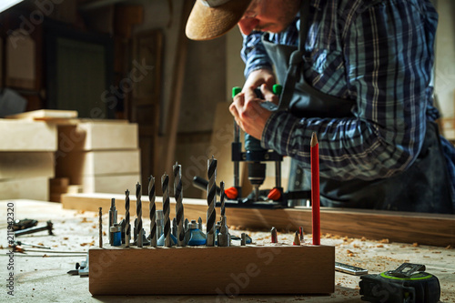 Experienced carpenter in work clothes and small buiness owner is carving a wooden board on an modern hand drill in a light workshop side view, in the background a lot of tools