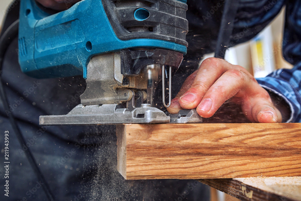 Close up of experienced carpenter in work clothes and small buiness owner  carpenter saw and processes the edges of a wooden bar with a jig saw  in a light workshop