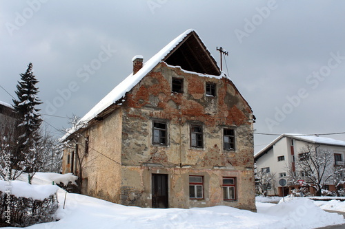 Abandoned old ruined house with broken windows, fallen facade and bricks from walls, covered in deep snow next to paved road and modern suburban house in background