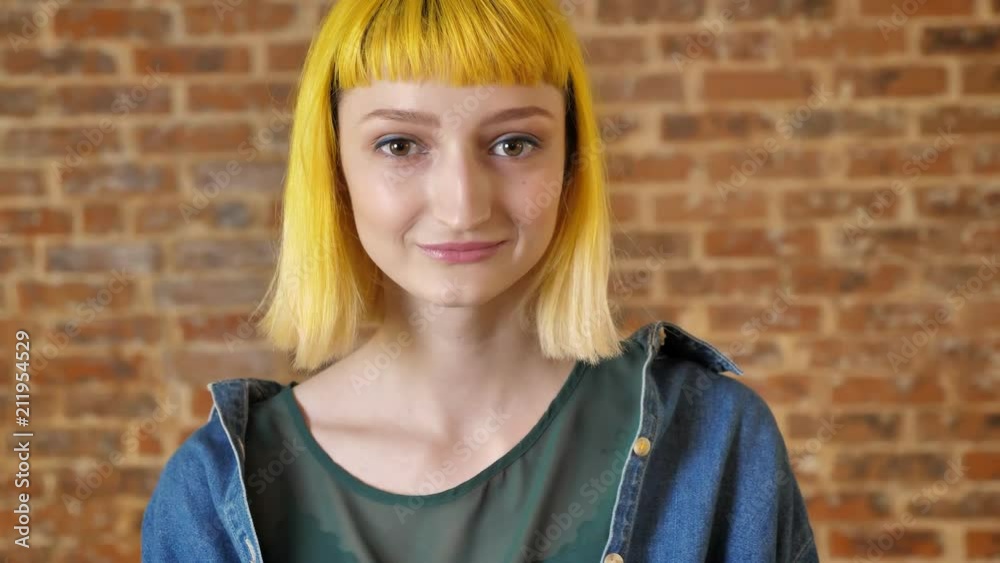 Young charming woman with yellow hair looking at camera and standing near brick background, smiling and positive Young charming woman with yellow hair looking at camera and standing near brick background, smiling and positive