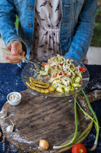 Woman hands holds raw vegan zucchini spaghetti with corn bread slices. Salad with red bell pepper, tomatoes, cucumbers and salad leaves dressed with sesame seeds and homemade tahini dip sauce paste. 