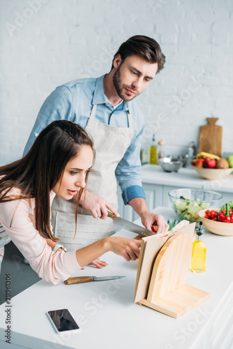 boyfriend cutting vegetables and girlfriend reading recipe in kitchen