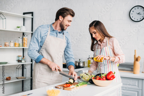 boyfriend cutting vegetables and girlfriend pouring oil into salad in kitchen