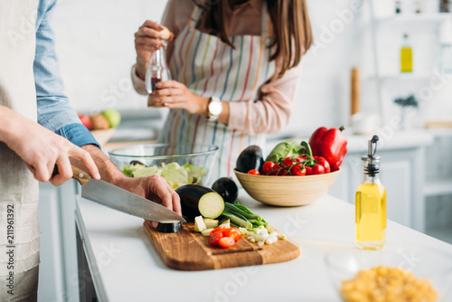 cropped image of boyfriend cutting ingredients and girlfriend adding spices to salad in kitchen