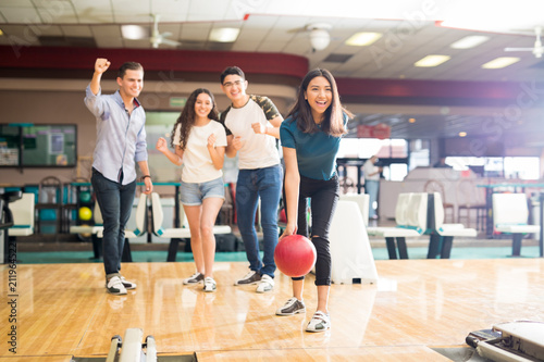 Friends Motivating Teen Girl Throwing Bowling Ball At Alley