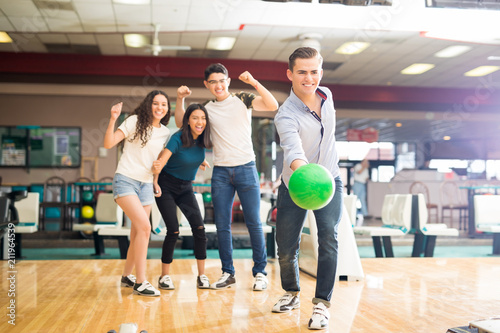 Boy Throwing Bowling Ball While Friends Cheering In Club