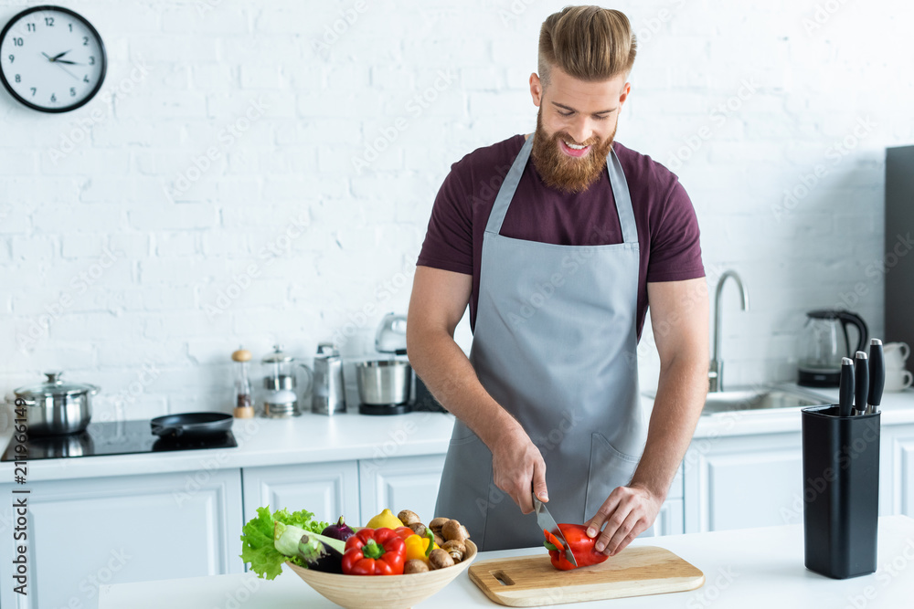 handsome smiling young man in apron cutting bell pepper on wooden cutting board handsome smiling young man in apron cutting bell pepper on wooden cutting board
