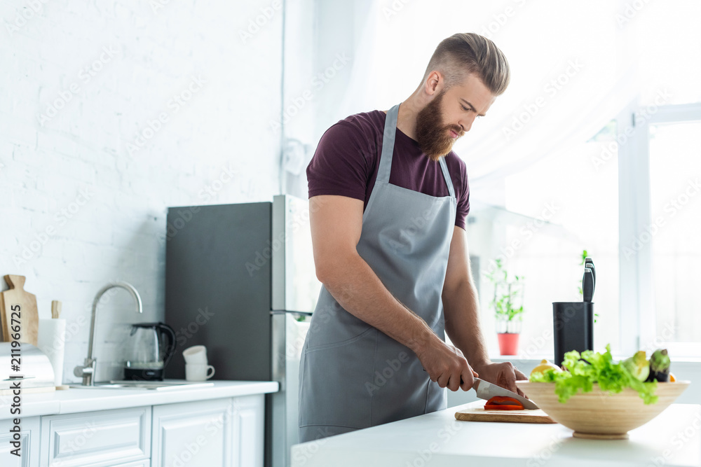 handsome bearded young man in apron cutting vegetables while cooking in kitchen handsome bearded young man in apron cutting vegetables while cooking in kitchen