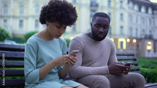 Girlfriend and boyfriend chatting in their smartphones, sitting on bench, ignore photo