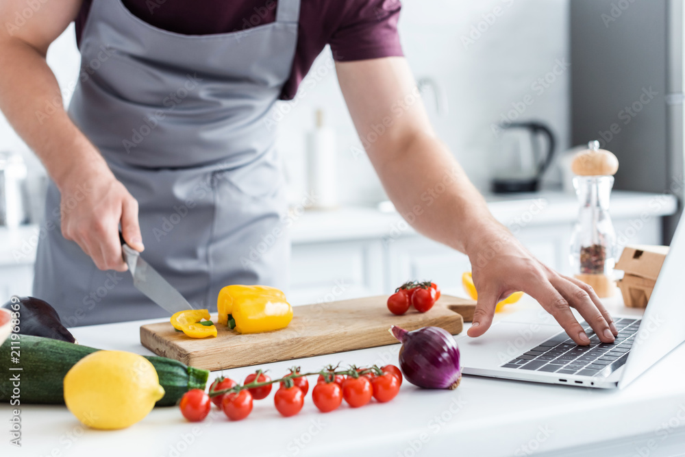 cropped shot of man in apron using laptop and cutting vegetables cropped shot of man in apron using laptop and cutting vegetables