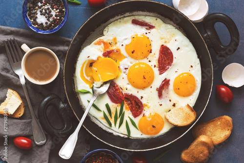 Flat lay fried eggs with tomatoes in a pan, toast with butter, coffee. A hearty Breakfast on a blue background.