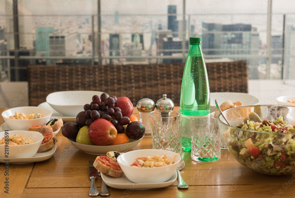Covered wooden table with food is on a background of window of a big city top view. 