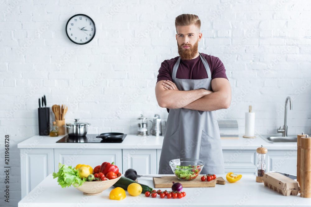 handsome bearded man in apron standing with crossed arms while cooking in kitchen