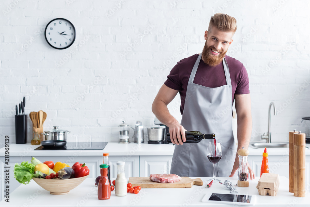 handsome bearded young man in apron pouring wine and smiling at camera while cooking in kitchen handsome bearded young man in apron pouring wine and smiling at camera while cooking in kitchen