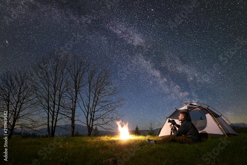 Beautiful night camping in mountains. Young male backpacker with photo camera sitting alone in front of tourist tent at burning campfire on grassy valley under night blue starry sky with Milky way