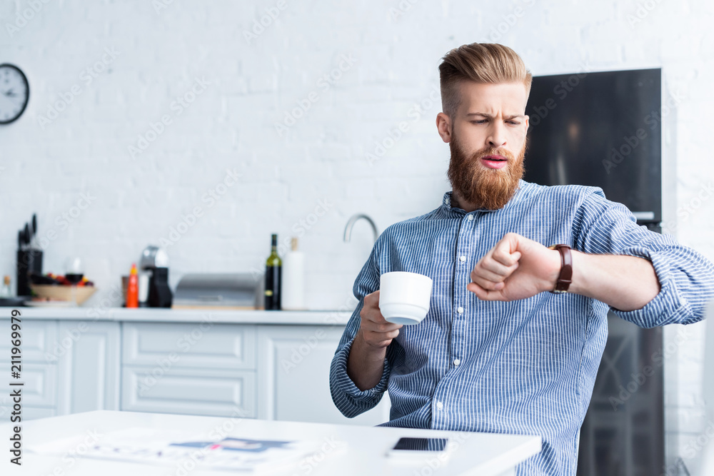 handsome bearded young man holding cup of coffee and checking wristwatch while sitting at table handsome bearded young man holding cup of coffee and checking wristwatch while sitting at table