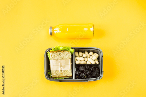 Lunchbox with food - sandwich, nuts and berries - next to a bottle of orange juice on a yellow background. Top view, flat lay,