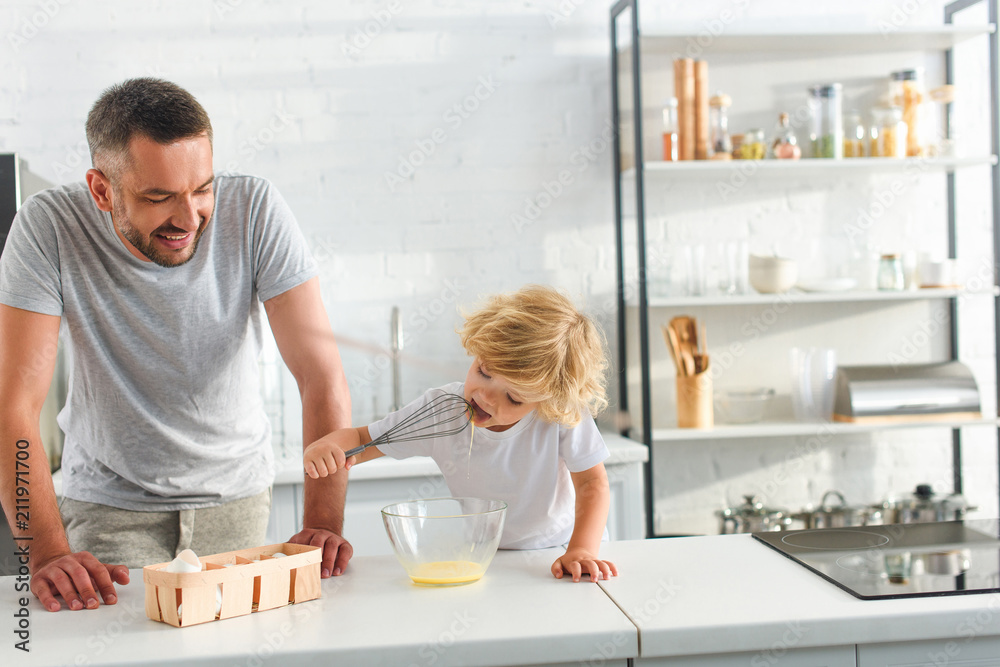 smiling father looking how his little son licking whisk with egg yolk at kitchen smiling father looking how his little son licking whisk with egg yolk at kitchen