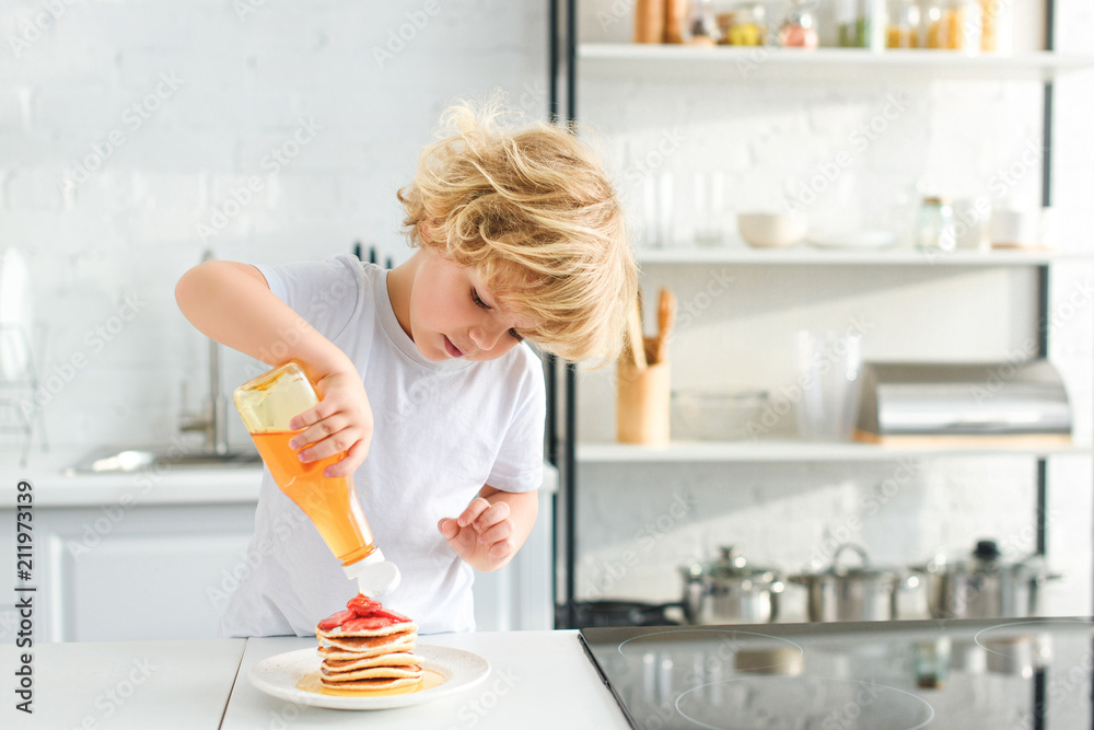 little boy pouring syrup on pancakes with pieces of strawberry on plate at kitchen