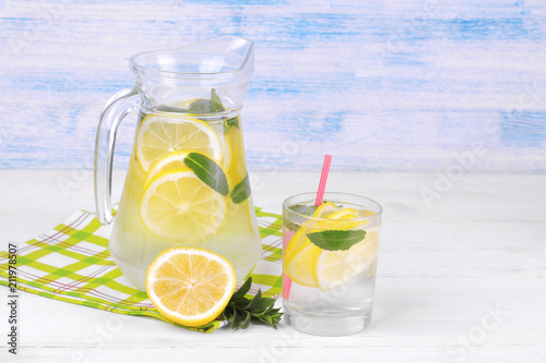 Homemade lemonade with lemon and mint in a glass jug and a glass next to fresh lemon on a white and blue wooden background
