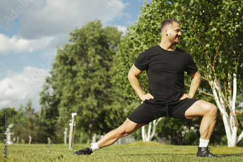 Young attractive man in black sportwear doing lunge outdoor in the park.